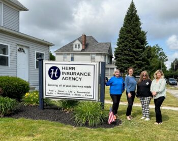 Katie and her team smile outside the Herrr Insurance Agency office, next to a large sign. 