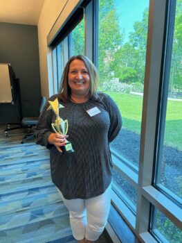 Michelle, a white woman with straight brown hair, a bright smile, a black and white patterned shirt and white capri pants, holds a gold trophy.