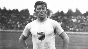 Jim Thorpe wears a white T-shirt with a shield with stars and stripes on his chest. He has an intense gaze and dark hair, short hair.