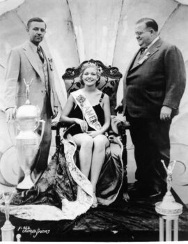 Marilyn smiles while wearing a tiara, sash and robe with two gentlemen on eithe rside of her. A large trophy is in the foreground.