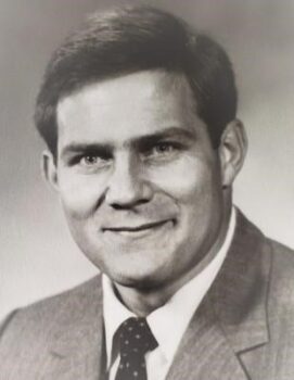 A black-and-white photo of a white man with short, dark hair, a smile, and a suit and tie. 