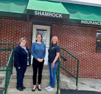 Katie and two Shamrock Insurance stand outside the brick building under a green awning.