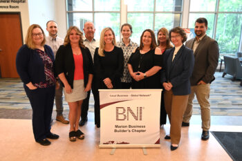 Members of the BNi group smile next to a sign with the BNI logo. There are about 11 men and women in profesional dress. 