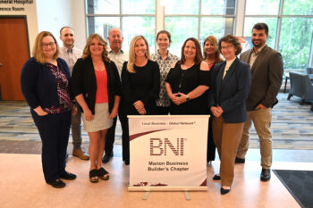 Members of the BNi group smile next to a sign with the BNI logo. There are about 11 men and women in profesional dress.