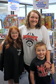 A white woman with shoulder-length curly hair smiles with her arms around two children. the white girl is as tall as her shoulder with long red, wavy hair and glasses. Teh Younger white boy has a buzz cut. 