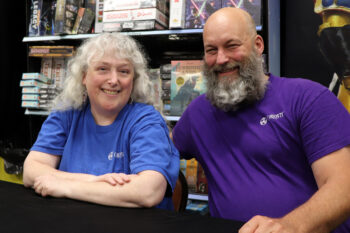 The owners lean into each other and smile. Jessica is a white woman with long, white curly hair and bangs and a blue T-shirt. Anthony is a taller white man with a bald head, gray and white beard covering his neck, and a purple T-shirt.