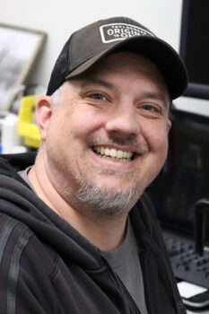 A white man with a smile, baseball hat, dark hoodie and goatee.