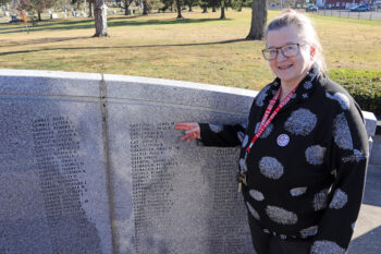 A white woman with light brown hair that is bpulled back, glasses, and a smile, points to names on the World War II memorial.