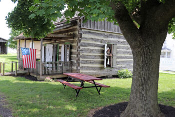 A volunteer with a beard looks out of a long window of a log cabin.It has wide weathered logs with white plaster in between. A small porch is visible on the front with an American flag. A building with light brickes is in the background.