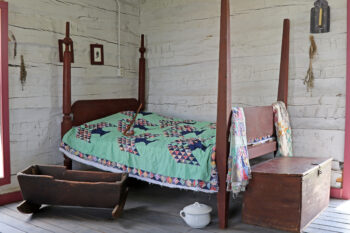 The inside of the cabin is made of logs painted white. A brown four poster bed is covered with a quilt. There is a wooden hope chest at the foot. A low wooden child's crib that can rock is next to the bed. A white chamber pot and lid is under the bed. Bundeles of lavendar, two pictures with silhouettes, and a scocen with a candle are on teh walls. 