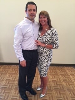 The couple stands and smiles. Bill is a taller man with short dark hair, a white buton-up shirt and black dress pants and shoes. Jac is a shorter woman with shoulder-length straight brown hair, a bright smile, a gold nexcklace, and a black and white patterned dress with silver heels.