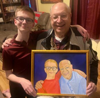 David Weichenthal, a senior with white hair, glasses, and a wide smile, poses next to his grandsonwith short blonde hair and glasses. They hold a painting of the two of them done by Dorothy Weichenthal