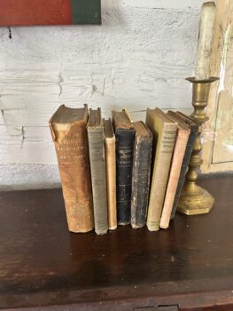 Old, well-worn books stand on a wooden desk next to a tall brass candle holder with a white candle. 