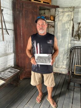 Jeff is a white man with a gray goatee wearing a baseball cap with sunglasses on the brim. He has khaki shorts and tan sandals. He holds a black and white photo showing him and other teens working to erect the cabin. 