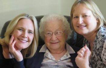 A mother is seated while her adult daughters lean in. Dorothy is a white woman with short, curled white hair, blue eyes and glasses with a delicate metal frame. She has a lavender, blue , and yellow patterned shirt with a sweater. Joan has blonde shoulder-length hair and a smile. Patty has blonde shoulderlenth hair and a smile.