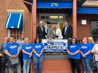 The employees stand in front of Laipply's. Six employees in blue shirts smile at the camera. On the porch, Jacqueline kisses her mother on the cheek. Their husbands stand next to them.