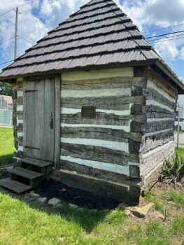 A square log cabin with white chink between the logs sits under a blue sky. It has a wooden door and three steps as well as a plaque. 