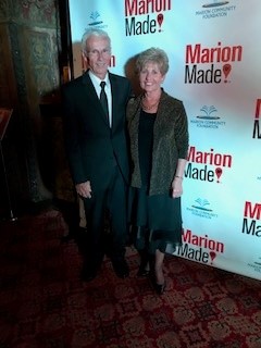 The couple stands in front of a MarionMade! backdrop. Ron is a taller man with short white hair, glasses and a smile. He wears a black suit with a white shirt and a black tie. Effie is shorter with dark blonde short hair, a black dress and a black jacket.