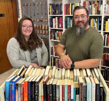 Crystal Bessler, owner of Birch Tree Bookery, stands with her husband Justin behind a library cart full of used books. Crystal is a white woman with glasses, a smile, long, wavy brown hair, a gray camo shirt and a gray patterned sweater. Justin is a taller white man with black hair with streaks of gray, a black and gray beard, glasses, an olive green patterned shirt, and a black Apple watch. Behind them is a wall of jewelery and more shelves of books.