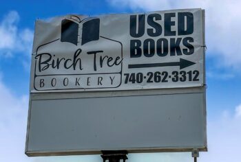 A white vinyl sign with the Birch Tree Bookery logo with a book with a birch tree pattern on the spine. The sign says Used Books with an arrow to the right and the phone number: 740-262-3312