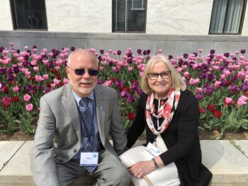 Gary is a white man with short white hair, a white beard, sunglasses, a gray suit, a blue shirt and a darker blue patterned tie. His wife is a white woman with shoulder-length blonde hair, black-rimmed glasses, a smile, a white dress with a black suit jacket and a pink and white scarf. 