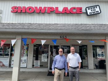 Two white men stand in front of a storefront with the Showplace Rent-to-own sign above them. The older man has short white hair and a beard, sunglasses, a blue button-down shirt, khaki pants and a black belt. The younger man is taller with a bald head, short red beard, a smile, a white button-downed shirt with short sleeves, gray slacks, a black belt, and a silver and black watch.