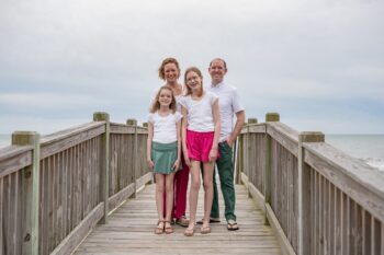 the family stands on a wooden bridge next to a beach and smiles. There are a woman, a man, and two girls. 