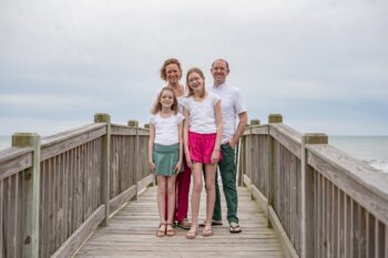 the family stands on a wooden bridge next to a beach and smiles. There are a woman, a man, and two girls.