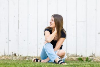 A senior picture of Emily. She is laughing while sitting in front of a white barn. She wears a black T-shirt and ripped, light, accid-wash jeans and black sandals. 