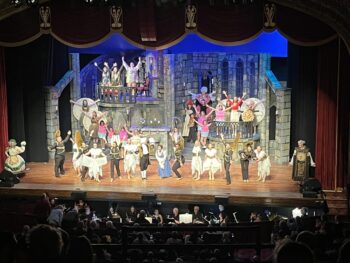 Cast members dressed up as furniture dance alongside Belle inside a castle.