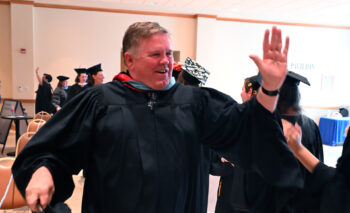 A white man with short brown hair in a black graduation gown high-fives students wearing gowns and mortar boards. 