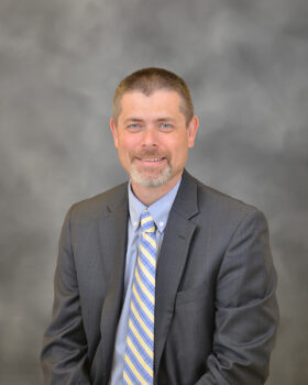 Ryan is a white man in his 40s with short brown hair, a gray beart, a gray suit, a light blue button-down shirt and a blue and yell striped tie. 