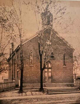 A two story brick building wtih a few narrow windows and a white bellow tower. A few trees are out front. 