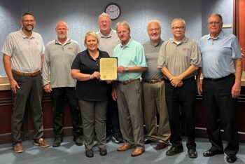 Commissioners and representatives of local companies smile while the resolution is displayed. There are seven white men and one white woman.