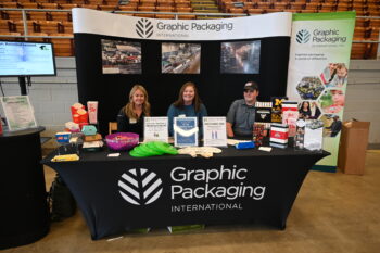 Three employees sit at a display for Graphic Packaging with boxes from Dairy Queen, McDonalds, Butter Finger, Popcorn, Panera Breads, Cracker Jack, and several sports brands.