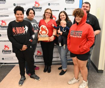 Elaine, a white woman with shoulder-length, brown wavy hair, glasses, and a wide smile, smiles while holding a wooden trophy with the Forge logo, a flame. She is surrounded by three adults, two teenage boys and one infant child. 