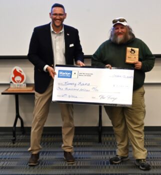 A white man presents a giant check to another white man with long gray hair, a white ball cap with sunglasses on the brim, a green sweatshirt and khaki pants.