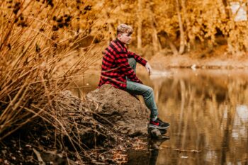 A young man with short light brown hair sits on a rock by a pond.