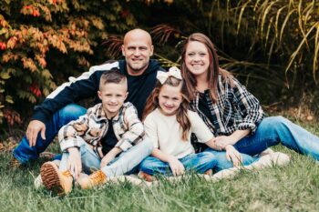 Two parents and their elementary age children sit on the grass and smile.