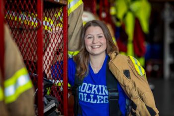 A teenager in a blue Hocking College T-shirt holds fire gear at the fire station and smles. 