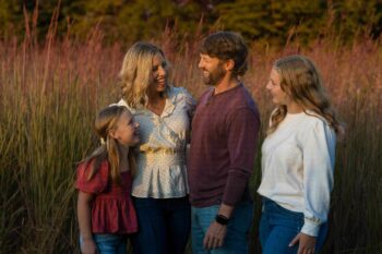 Two parents smile and laugh with their daughters in front of tall grass.