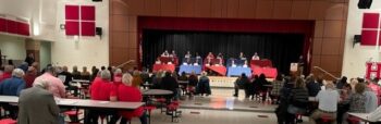 Candidates behind tables on a stage answer questions in front of a crowd of voters.
