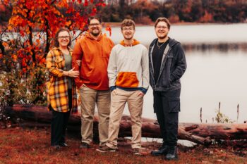 The family poses and smiles in front of a pond with fall lfeaves behind them.