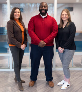 Two white women stand on either side of a tall black man with a beard.