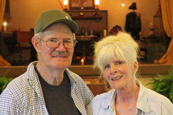 A white man with glasses, white short hair, a white and brown mustache, a T-shirt and plaid collared shirt over top has his arm around his wife. Donna is a white woman, slightly shorter, with long white hair up in a bun and bangs. She smiles at the camera. She has a light demin shirt with a collar.