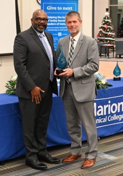 A tall bald black man with black-rimmed glasses and a short beard smiles while receiving a blue glass trophy from a white man with salt-and-pepper hair and a short gray beard. 