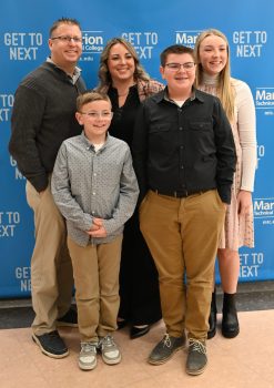 Natalie, a white woman with light brown curly hair, smiles while standing next to her husband and three children. 