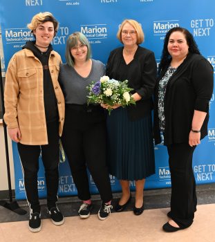 Darlene Schoonard, a tall white woman with a dark blonde bob and glasses, smiles while holding a bouquet of flowers. She is surrounded by her friends and supporters: two young woman and a young man. 