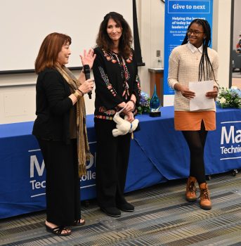 Trish, a tall white woman with long brown hair, smiles as Lillie, a shorter white woman with reddish-brown hair, and Jessica, a young Black woman with long black braids and glasses, induct her.