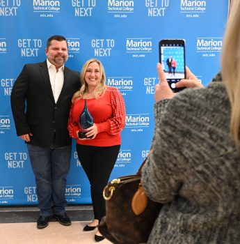 A short white woman with a smile and long blonde hair holds her trophy while posing next to her husband, a white man with dark short hair, a gray short beard. Blair's iphone and hand and blonde hari can be seen as she takes a photo. Blair nominated her employee for the Marion Technical College Alumni Hall of Fame.